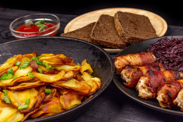 Roasted potato in the pot, pieces of rye black bread on a wooden serving plate, homemade sausages in bacon and stewed cabbage on a black rustic background