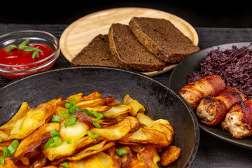 Roasted potato in the pot, pieces of rye black bread on a wooden serving plate, homemade sausages in bacon and stewed cabbage on a black rustic background
