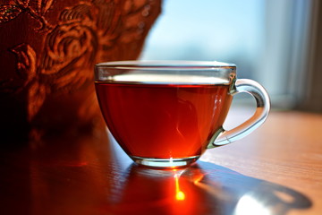 Glass cup of tea on wooden window sill in front of a dark curtain in sunrise on a blurred background