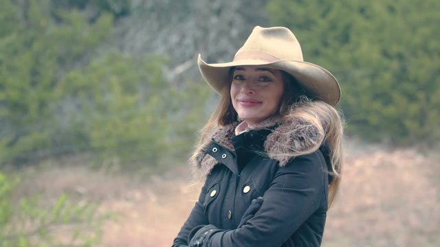 A smiling cowgirl stands outside in the cold then looks down and is deep in thought.