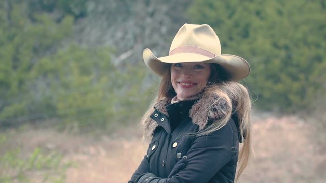 A woman wearing a cowboy hat stands alone with crossed arms, smiles flirtatiously.