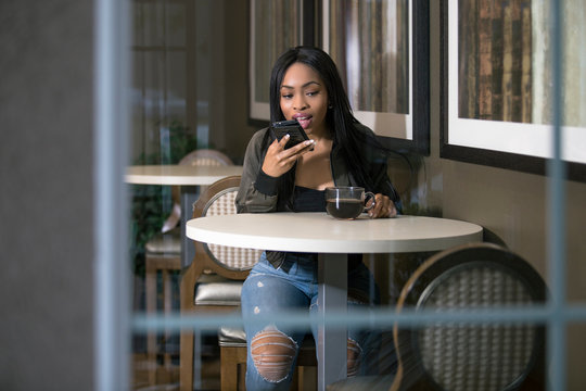 Window view of a black female using a mobile cellphone in a coffeeshop or sidewalk cafe.  She is sitting and having coffee while holding a smart phone.