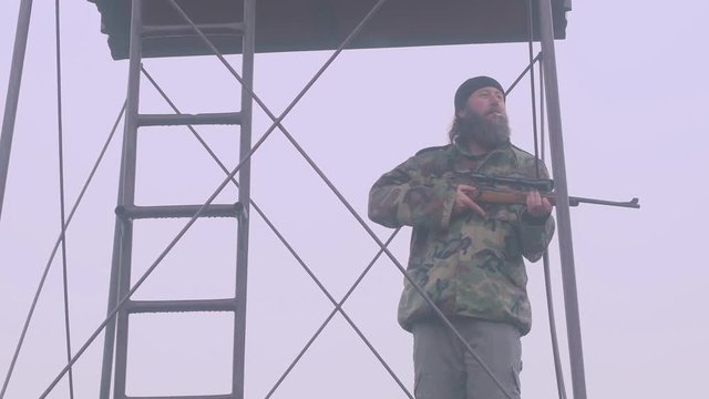 Man guards tower with sniper rifle, standing alone smoking on alert