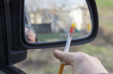 close-up, a man's hand holding a Smoking cigarette in the car window, which stands in the courtyard of a residential area overlooking a children's Playground in the rear view mirror
