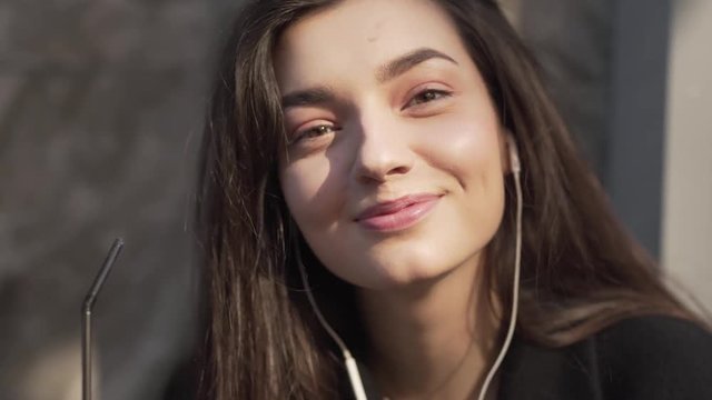Closeup of a beautiful young woman listening to music through earphones and sipping a beverage from a straw while looking at the camera and smiling.