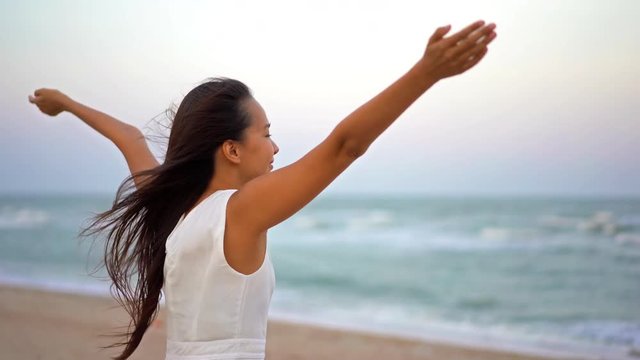 Asian Female in White Dress and Long Hair Standing By Sea on the Beach With Hands in Air Looking at Sea on Windy Afternoon