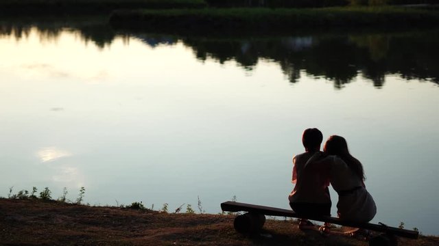 Backside view of two girls sitting on a bench by peaceful lake with reflections from trees in the golden hour