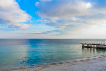 Contrast photograph of the sea and cloudy sky. In the water is the construction of the pier, which leads to the sandy shore