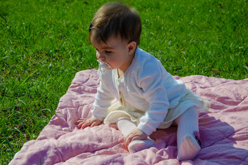 Baby in bright clothes on a pink plaid on green grass in the park. Newborn baby outside on a warm day. Little girl plays on a plaid on a green lawn in spring