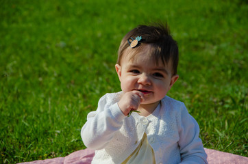 Fototapeta premium Baby in bright clothes on a pink plaid on green grass in the park. Newborn baby outside on a warm day. Little girl plays on a plaid on a green lawn in spring