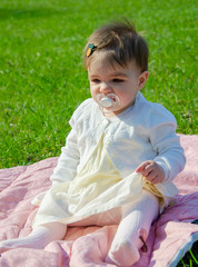 Baby in bright clothes on a pink plaid on green grass in the park. Newborn baby outside on a warm day. Little girl plays on a plaid on a green lawn in spring