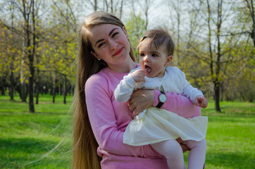 Fototapeta premium Mom with baby in bright clothes on a pink plaid on the green right. Family resting in the park on a warm day. Mom and little girl 10 months walk in the park