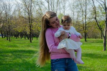 Mom with baby in bright clothes on a pink plaid on the green right. Family resting in the park on a warm day. Mom and little girl 10 months walk in the park