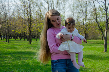 Fototapeta premium Mom with baby in bright clothes on a pink plaid on the green right. Family resting in the park on a warm day. Mom and little girl 10 months walk in the park