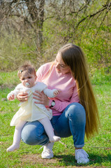 Fototapeta premium Mom with baby in bright clothes on a pink plaid on the green right. Family resting in the park on a warm day. Mom and little girl 10 months walk in the park