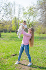 Fototapeta premium Mom with baby in bright clothes on a pink plaid on the green right. Family resting in the park on a warm day. Mom and little girl 10 months walk in the park
