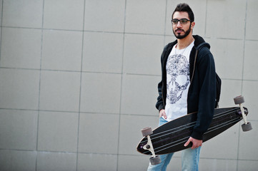 Street style arab man in eyeglasses with longboard posed against gray wall.