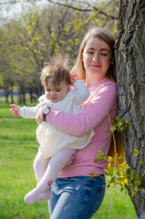Fototapeta premium Mom with baby in bright clothes on a pink plaid on the green right. Family resting in the park on a warm day. Mom and little girl 10 months walk in the park