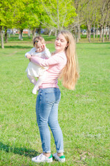 Fototapeta premium Mom with baby in bright clothes on a pink plaid on the green right. Family resting in the park on a warm day. Mom and little girl 10 months walk in the park
