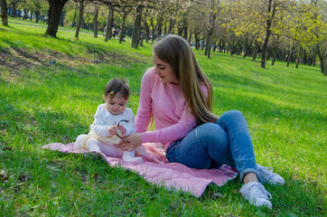 Fototapeta premium Mom with baby in bright clothes on a pink plaid on the green right. Family resting in the park on a warm day. Mom and little girl 10 months walk in the park