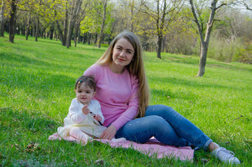 Naklejka na ściany i meble Mom with baby in bright clothes on a pink plaid on the green right. Family resting in the park on a warm day. Mom and little girl 10 months walk in the park