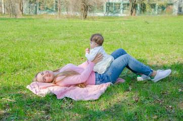 Mom with baby in bright clothes on a pink plaid on the green right. Family resting in the park on a warm day. Mom and little girl 10 months walk in the park