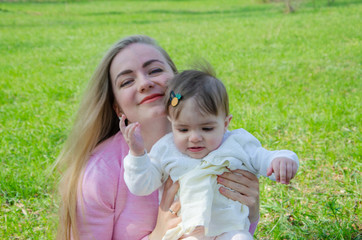 Fototapeta premium Mom with baby in bright clothes on a pink plaid on the green right. Family resting in the park on a warm day. Mom and little girl 10 months walk in the park