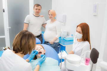 Fototapeta premium A female dentist and her assistant are treating the teeth of a little girl. Parents of the girl oversee the treatment. Mom girl is pregnant