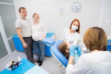 A female dentist and her assistant are treating the teeth of a little girl. Parents of the girl oversee the treatment. Mom girl is pregnant