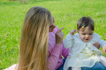 Mom with baby in bright clothes on a pink plaid on the green right. Family resting in the park on a warm day. Mom and little girl 10 months walk in the park