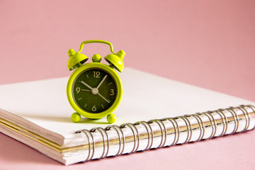 A small green alarm clock is standing on a white notepad on a pink background. The clock shows the time of 10 am. Background with a clock. Time work. Copy space