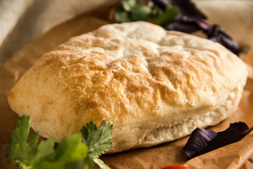 Ciabatta italian white bread on parchment paper on a wooden table, next to fresh greens and cotton napkin.