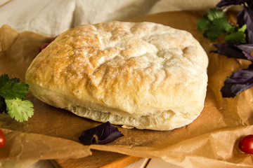Ciabatta italian white bread on parchment paper on a wooden table, next to fresh greens and cotton napkin.