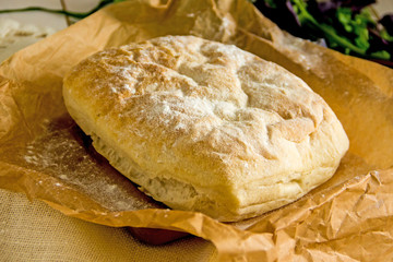 Ciabatta italian white bread on parchment paper on a wooden table, next to fresh greens and cotton napkin.