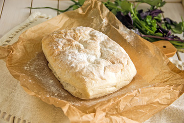 Ciabatta italian white bread on parchment paper on a wooden table, next to fresh greens and cotton napkin.