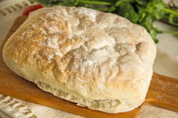 Ciabatta italian white bread on parchment paper on a wooden table, next to fresh greens and cotton napkin. View from above