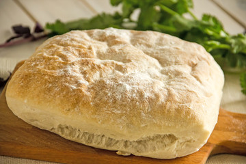 Ciabatta italian white bread on parchment paper on a wooden table, next to fresh greens and cotton napkin.