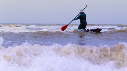 A woman with a cap on the head, practicing stand up paddle, on the North Sea in the Netherlands with large waves and strong winds.