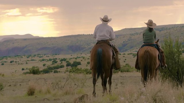 Vintage style, husband and wife ranchers watch the sunrise on horseback, Rocky mountain range ahead, 29 fps.