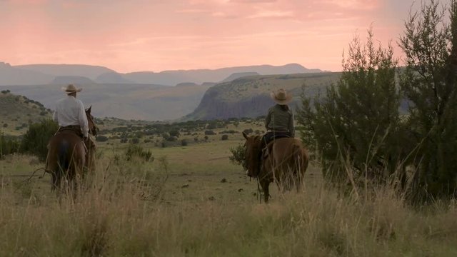 Cowboy and cowgirl ride horseback into a dramatic pink sky and mountain range backdrop, slow motion 24 fps.
