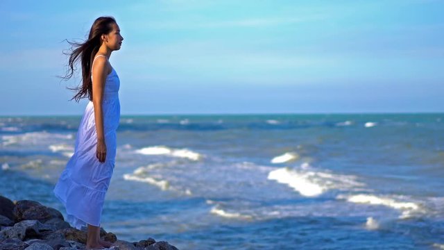 Asian woman with long hair and white dress stands looking over ocean on windy day