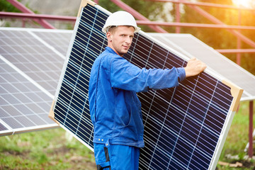 Young worker in blue uniform and protective helmet carrying big shiny solar photo voltaic panel to...