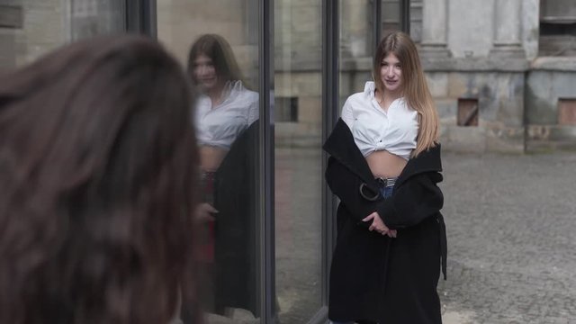 Young Female in White Shirt Jean Pants and Black Coat Posing by Glass Wall While Her Friend Taking Pictures With Digital Camera and Pointing Where She Have to Stand. Light Camera Movement