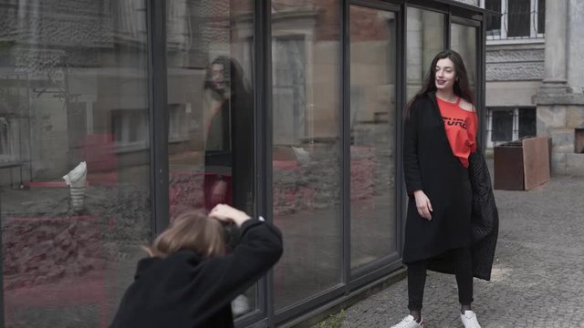 Teenage Girl in Red Shirt and Black Coat Walking by Glass Wall and Posing While Her Friend with Brown Hair Taking Pictures of Her With Digital Camera. Blurred Background