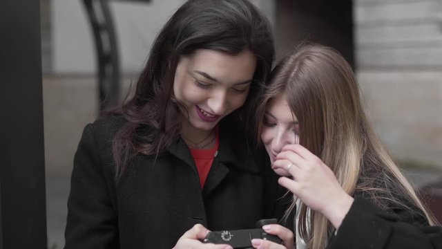 Young Female in Black Hair and Coat Holding Digital Camera and Looking Images on Display With Her Brown Haired Friend Who is Huging Her and They Talk. Light Camera Movement