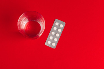 Pills in blisters with glass of water on red background top view. Medicine