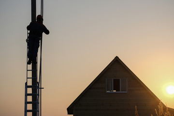 electrician performs installation of electrical equipment on the power line support near a wooden house in the evening at sunset