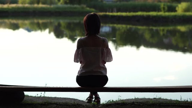 Backside view of a woman sitting on a bench in summer clothing on peaceful lake background with lush green tree reflections in water