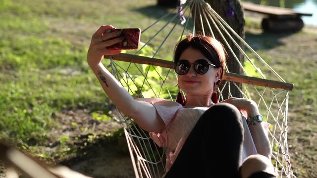 Frontal view of young woman in pink blouse and sunglasses relaxing and  taking a selfie in mesh hammock on a green grass background