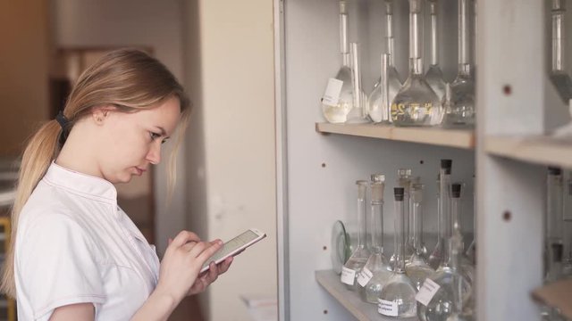 Young charming female laboratory worker is counting some empty flasks and write it down in a phone. She is standing in a pharmaceutical laboratory.